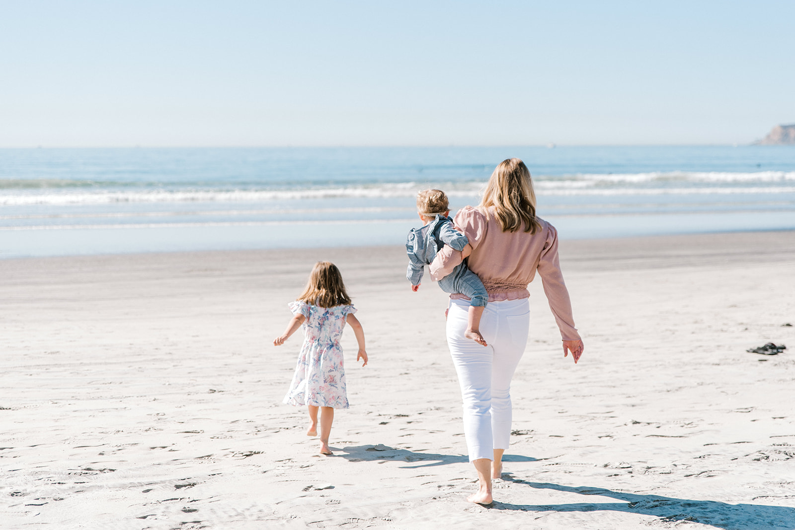 Woman in white pants walks toward the ocean carrying a toddler as a young girl walks ahead during Coronado Beach Family Photos