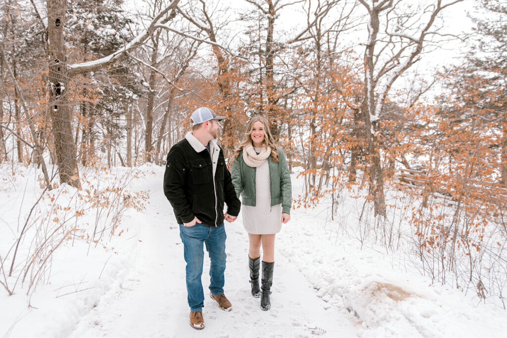 A woman in cream dress and scarf with green jacket walks in the woods with her fiance during a winter engagement session