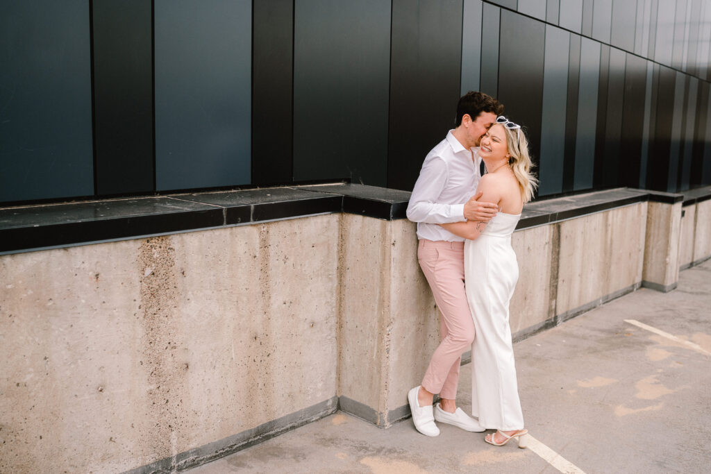 A woman in a white jumpsuit with white heart sunglasses smiles as a man whispers in her ear during an engagement photo session