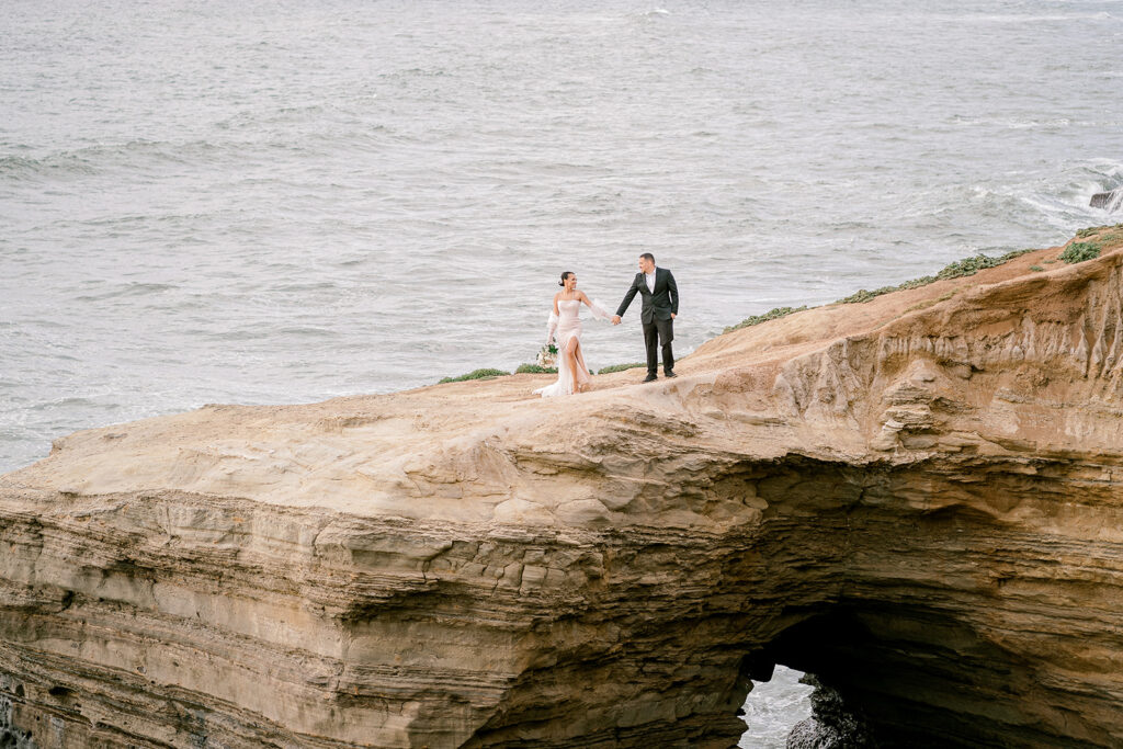 A bride and groom hold hands and look at each other on a cliff overlooking the ocean at Sunset Cliffs in San Diego, California. 