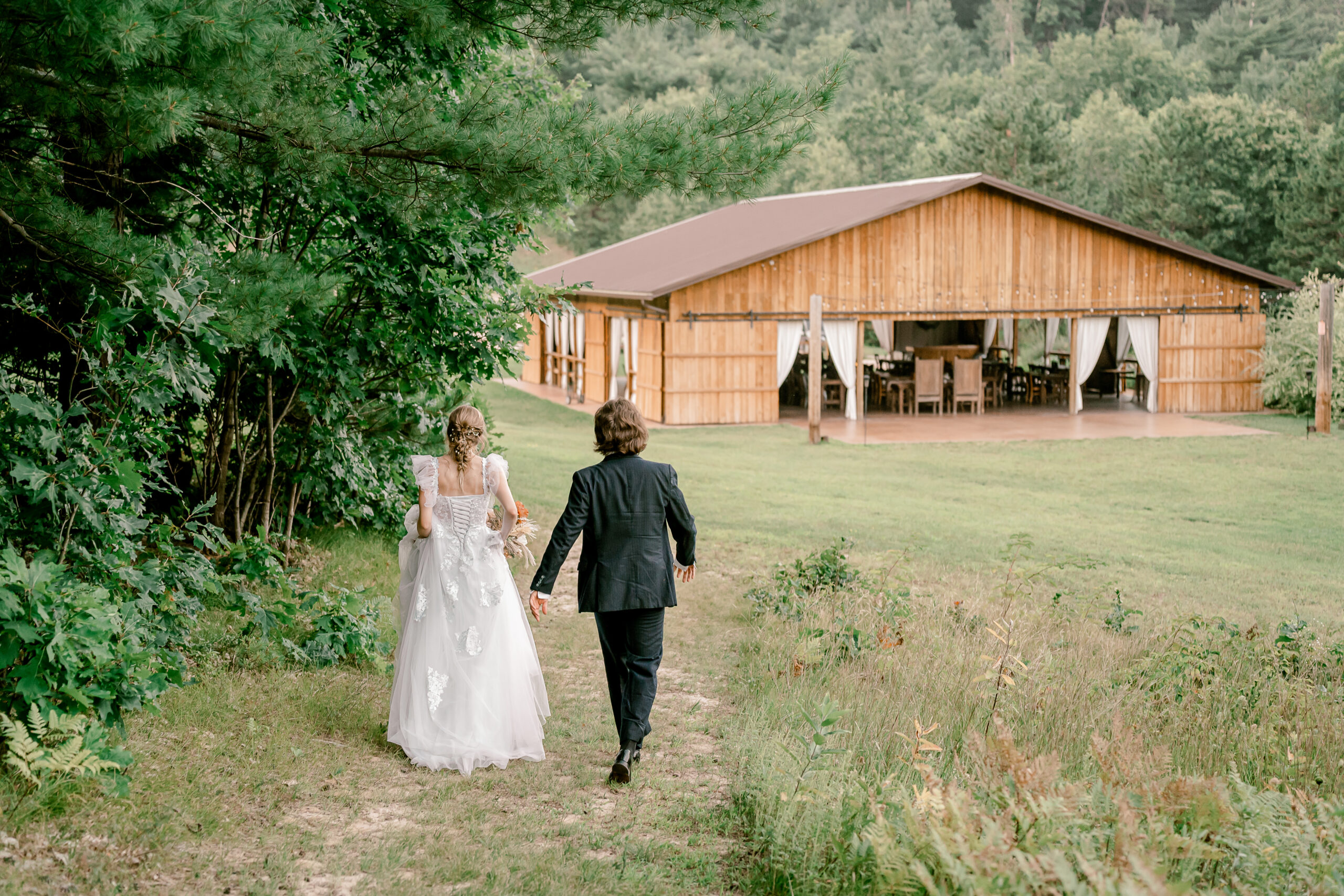 A bride and groom walk along a wooded path to the outdoor pavilion at Burlap and Bells