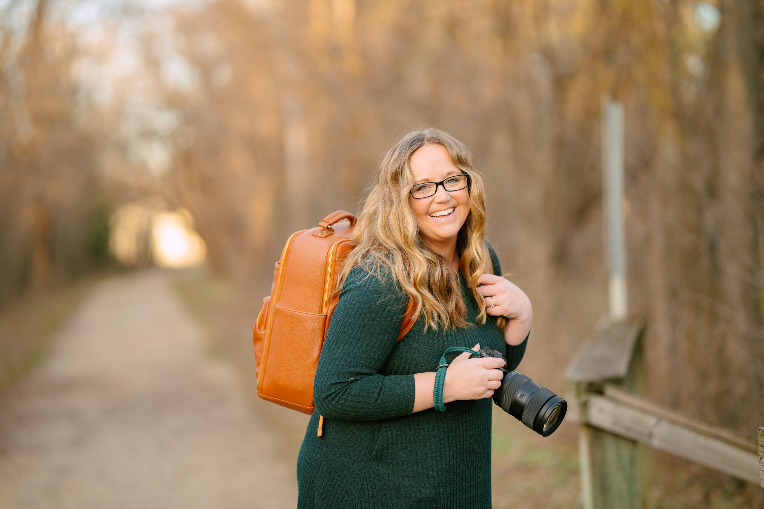 A woman in a green dress carries a camera and wears a caramel colored backpack
