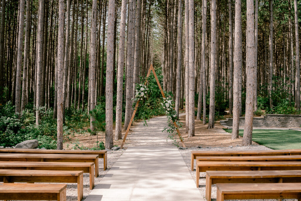 Wooden Benches line the aisle at the ceremony location at Pinewood.  A triangle arch is adored with white flowers and greenery. 