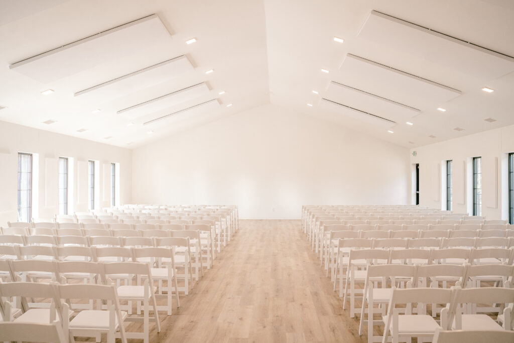 The indoor ceremony space at Pinewood Weddings and Events.  Gorgeous white walls with a lot of natural light coming in from the windows and white chairs in rows for a wedding ceremony. 