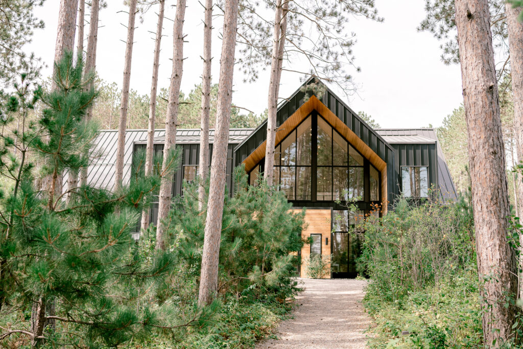 A path through wooded pine trees leads to a modern black building with large windows at Pinewood Weddings and Events