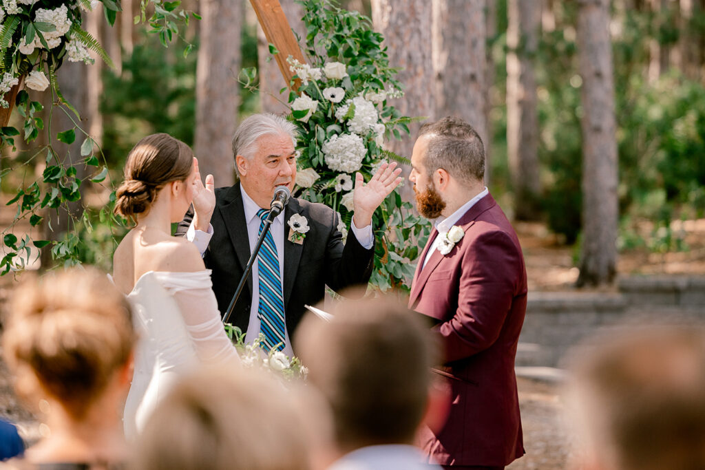 a couple is at the alter and their officiant is speaking to them with his hands in the air.  He tells guests to put their phones away for an unplugged wedding ceremony. 