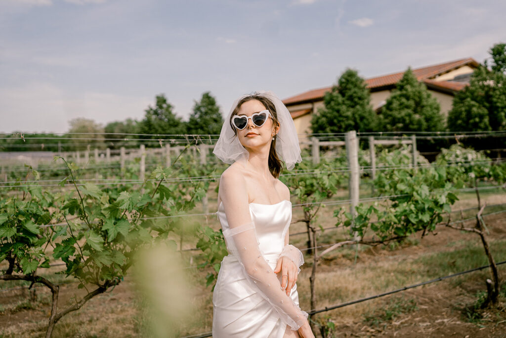 A bride in a veil and sleeves with pearls wears white heart glasses while in a vineyard on a sunny day