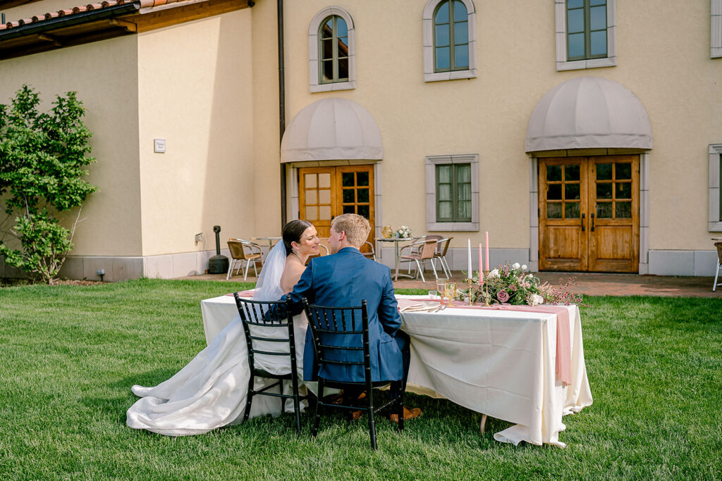 A bride and groom share an intimate moment while seated at a table at their Villa Bellezza wedding.  The stucco building with terracotta roof is in the background. The table has a pink and green bouquet on it. 