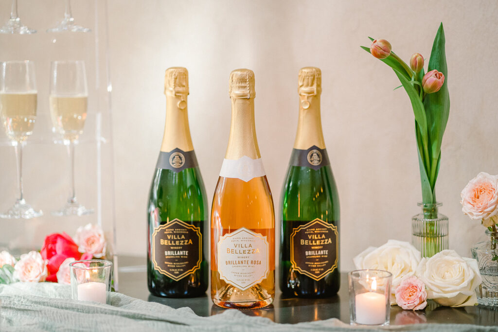 Three bottles of Villa Bellezza champagne sit on a table, surrounded by candles, flowers and champagne glasses. 