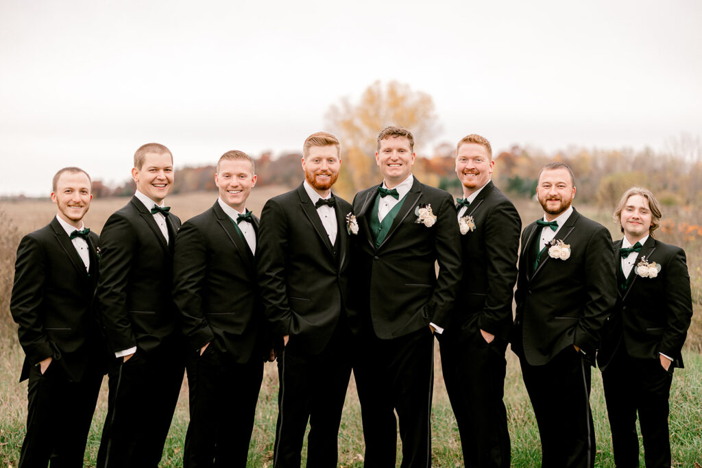 A groom and his groomsmen stand in a field in black tuxes and smile at the camera