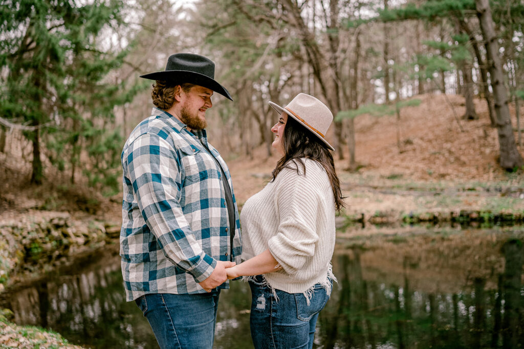 engaged couple smiling at each other while holding hands in front of a pond at rod and gun park during their engagement session