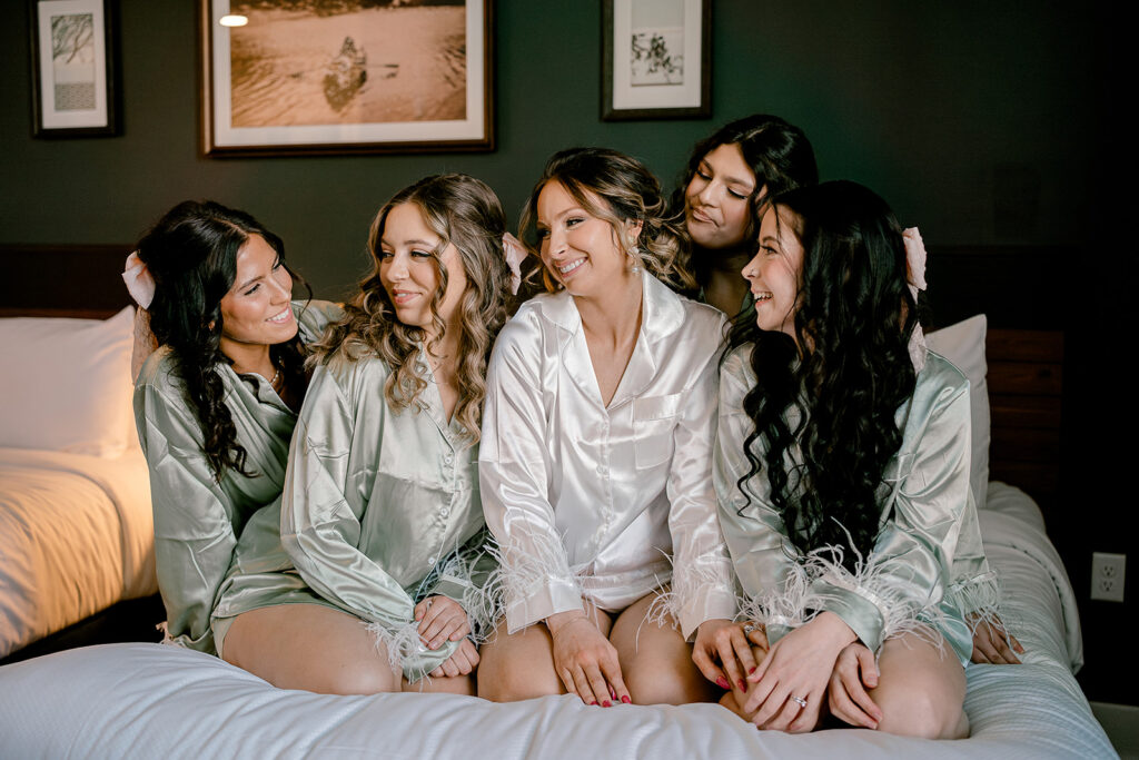 A bride and her bridesmaids in cute satin robes before her wedding day. They are on a bed, looking at each other. 