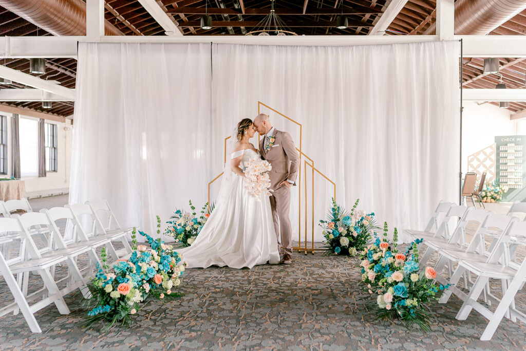 White chairs line the aisle in a ballroom. A bride and groom stand forehead to forehead at the end of the aisle.