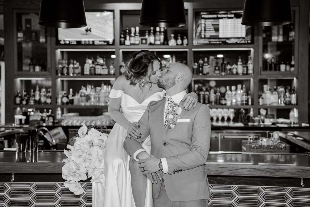 A couple in wedding attire kisses. The bride sits on a bar at the Confluence Hotel and her groom stands next to her. 