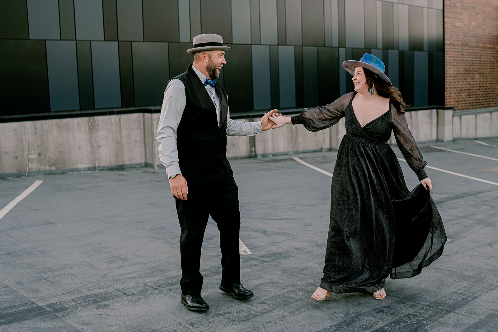 A woman in a dress and a blue hat dresses with a man in a vest and hat on a rooftop during their engagement photo session